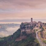 The "dying city" Civita di Bagnoregio rises on a rocky outcrop with pink skies at sunrise, mist lingers in the valleys around the ancient village