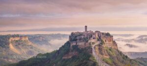 The "dying city" Civita di Bagnoregio rises on a rocky outcrop with pink skies at sunrise, mist lingers in the valleys around the ancient village