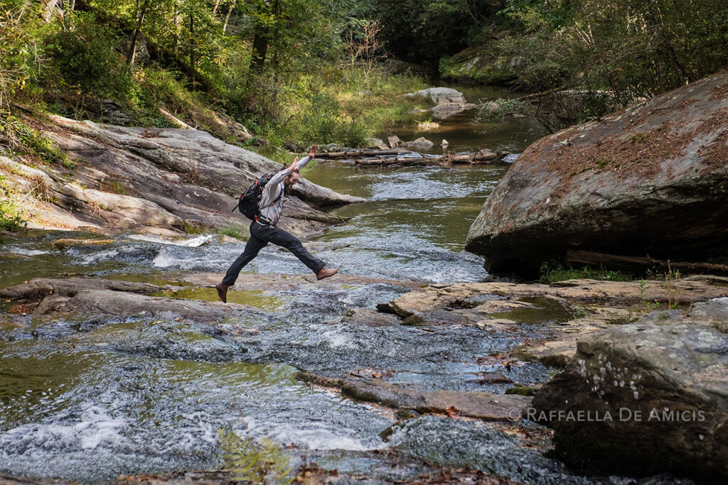 A hiker leaps across rocks in a shallow river while hiking in the north Georgia mountains, USA