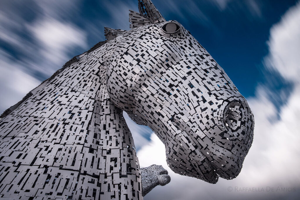 the massive metal statue of the kelpies (a horse head) in Scotland looms with blurred clouds in the background