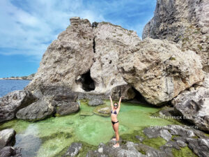 woman in a bikini poses with hands in air and big smile on rocky edge of an ocean pool in the Tremiti Islands of Italy