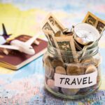 jar of currency labeled travel sits on map with toy plane and passport in background for article about travel budget mistakes to avoid