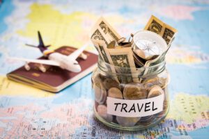 jar of currency labeled travel sits on map with toy plane and passport in background for article about travel budget mistakes to avoid